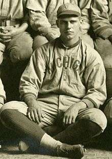 A man, wearing a grey baseball cap, baseball jersey with "MICHIGAN" on the chest and dark baseball socks, sits on the floor with his legs crossed.
