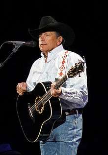 A middle-aged man in a white shirt and a black cowboy hat playing a guitar and singing into a microphone