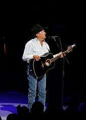 A man wearing a black cowboy hat, white shirt and blue jeans, playing a guitar and singing into a microphone