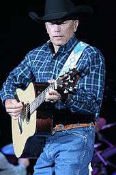 A middle-aged man in a black cowboy hat, checked shirt and jeans, playing a guitar