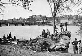 Eleven soldiers idly lining the shore of a river. On the opposite bank, connected by a bridge, are short buildings along the river and on top of a hill.
