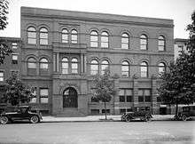 A rectangular, Victorian-style brick building with several trees and three cars from circa 1920 in front.