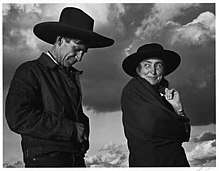 A black and white photograph shows Georgia O'Keeffe and Orville Cox wearing hats with the sky and clouds behind them.