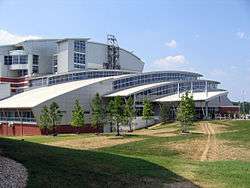 A large, white, multi-story building constructed from concrete, metal and glass with several tiered, curved roof segments framing long panels of windows. The building is set back on a large green lawn with several small pine trees