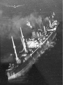 Black and white aerial photo of a merchant ship with splashes in the water along her port side. A monoplane aircraft is flying just above the ship.