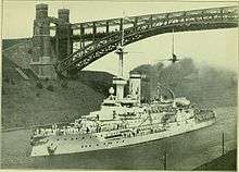 A large warship passing through a narrow canal, with a large railway arch bridge above it