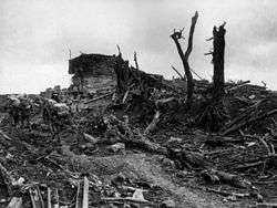 Heavily laden soldiers trudge through mud past the shattered remains of a concrete structure. Around them broken trees, steel beams and other pieces of debris have been strewn.