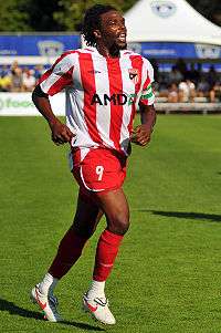 A black man with mid-length hair and a beard jogs across a grassy surface.  He is wearing a red and white striped soccer jersey, red shorts and red socks.