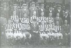 A football team comprising ten players in striped shirts and one in a shirt of a single colour pose for the camera. Five of the men are standing and the other six seated in front of them. Also posing with them are an elderly man in a bowler hat with a chain of office around his neck, and twelve men in business suits, some of whom are wearing hats.  A crowd of spectators is visible behind the group.