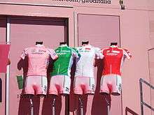 Four cycling jerseys&nbsp;– pink, green, white, and red, from left to right&nbsp;– are arranged on mannequin torsos in front of a pink wall.