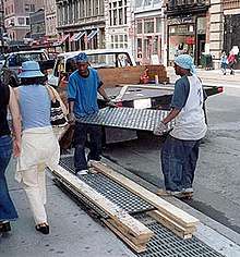 Two people in work gloves carefully transferring a heavy panel of pavement lights from a small flat-bed truck to a pile of similar panels, separated by wooden spacers. The pile is on the outside edge of the sidewalk, and pedestrians pass them on the inside. The panel has a black metal frame and round white lenses. It is about 1.5x0.5m (possibly 2x4 feet)