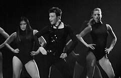 The grayscale picture of three people, two women and a man, who dance and put their hands on their hips. The man wears a dark outfit, compound of a vest, a shirt and pants. The women wear similar leotards.
