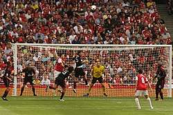 A colour photograph of an incident in the Milan penalty box at the Emirates Stadium; there are two Arsenal players in view and hundreds of home spectators in the background. As the ball is entering the box, Milan players are positioning themselves to defend.