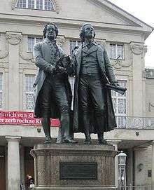 Photograph of a large bronze statue of two men standing hand-in-hand, side-by-side and facing forward. The statue is on a stone pedestal, which has a plaque that reads "Dem Dichterpaar/Goethe und Schiller/das Vaterland".