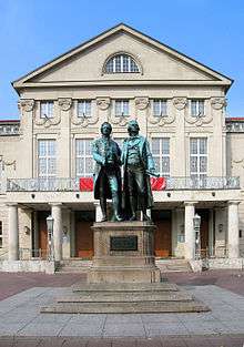 Photograph of a large bronze statue of two men standing side-by-side and facing forward. The statue is on a stone pedestal, which has a plaque that reads "Dem Dichterpaar/Goethe und Schiller/das Vaterland". Behind the monument there is a large, 3-storey building with an elaborate stone façade.