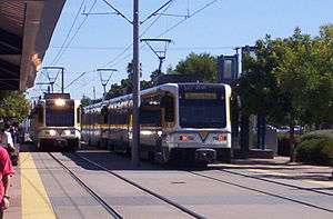 A pair white and yellow light rail trains are visible, with overhead lines above, and several passengers visible on the station platform.