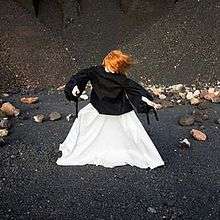 A red headed woman wearing a black shirt and white skirt stands in the middle of a black desert with the wind blowing her arms and her hair to the right side of the cover.