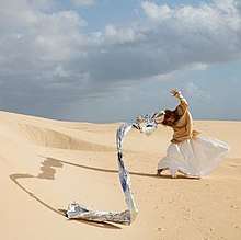 A red headed woman wearing a beige shirt and white skirt stands in the middle of a desert with the wind blowing her arms and a large drapery that flows from her hand.