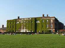 Massive, rectangular, three-storey brick building, covered in ivy. People are sitting in groups on the large front lawn.