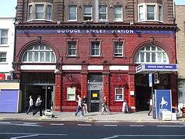 A red-bricked building with a blue sign reading "GOODGE STREET STATION" in white letters and several people walking in front all under a blue sky