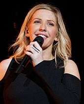 A smiling blonde woman with a black top holding a microphone.