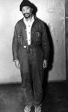 A black African man wearing overalls looks seriously at the camera.