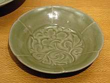 A green-grey plate with a leafy vine pattern painted into the center. The edge is divided into six sections, each arched slightly outward, to create the illusion that they were flower petals.