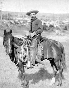 A black or white photograph of a cowboy posing on a horse with a lasso and rifle visible attached to the saddle