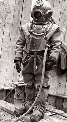 Standing figure of a diver clad in copper helmet, heavy canvas diving suit, with gloves, chest weight and weighted boots