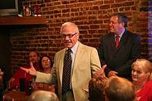 Barr stands in front of a brick wall in a vanilla suit, with his arms stretched to convey a point, before the small crowd around him