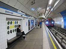 An underground station platform with white tiled walls, a white panelled ceiling arching over the track. The tiled a recess behind a platform seat features a design of twelve coloured circles arranged in a 4 by three grid; mostly dark green, but with three yellow, two black and one blue circles.