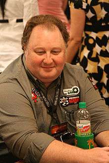 image of Greg Raymer seated at the 2008 World Series of Poker, wearing a green shirt with player patches applied and its long sleeves folded up to his elbows, with a black lanyard and badge around his neck