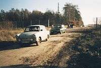 An East German Trabant and a Lada driving along a dirt track through a gap in the border fence. A border guard is visible by the side of one car, and a group of pedestrians can be seen in the background standing by the side of the road as it runs along the edge of a forest.