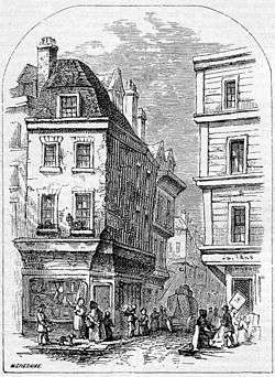 People congregate at the entrance to a narrow street, overlooked by two four-storey buildings.  Each floor of the right-most building projects further over the street than the floor below.  At the corner of each building, shops advertise their wares. A cart is visible down the street, and one man appears to be carrying a large leg of meat.