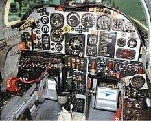 Aircraft cockpit with numerous old circular dials and gauges. In front of the controls is a black stick control column.