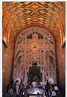 Interior photograph of high, arched ceilings, elaborately patterned with brightly colored designs, while an intricate metal screen partially hides a large mural behind.