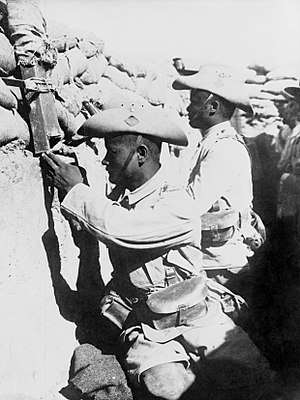 Two Gurkha soldiers in full uniform with turned-up slouch hats sit on the fire step in a fighting trench topped with sandbags. One soldier is looking through a periscope.