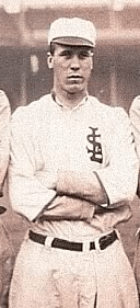 A man in a white baseball uniform with an overlapping "STL" on the left breast stands on a baseball field with his arms crossed.
