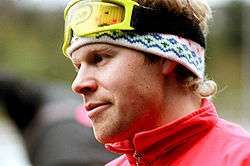 A blond man wears a red winter sports jacket and yellow ski goggles over a white wool head band.