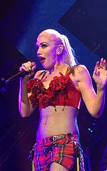 A blonde lady sings into a microphone, which is held in her right hand, in front of a blue background. She is wearing a red top and plaid shorts.