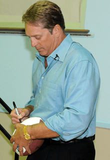 Color full-length photograph of stocky, well-tanned white man (Jack Del Rio), wearing teal blue button-up shirt and black pants while signing a football.