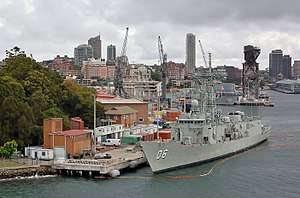 Colour photo of two grey-painted warships moored alongside wharfs. A large crane and several buildings are visible behind the ships.