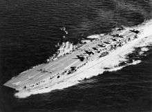 Aerial view of an aircraft carrier travelling at speed. Multiple personnel and aircraft are on the flight deck.