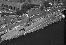 Aerial photograph taken from directly overhead of a small aircraft carrier, while she is berthed alongside a wharf. Nine aircraft are parked in a three-by-three pattern at the carrier's stern, and personnel are positioned to spell out the word "Aloha" when seen from above.
