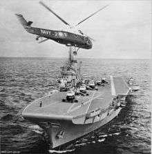 A small aircraft carrier is sailing towards the frame, with five helicopters in a line on the carrier's deck. A sixth helicopter hovers in the foreground