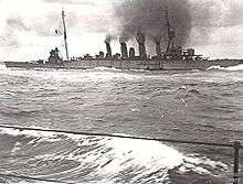 A large four-stacked warship billowing thick black smoke and moving through moderate seas.