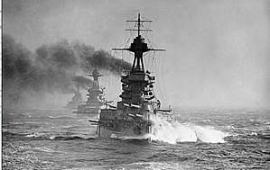 A large gray battleship steams in choppy seas; thick black smoke billows from its funnels. Two battleships are directly behind