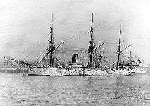 Broadside view of a metal ship, quiet at anchor in a port. Two small boats are alongside. There are three masts but no sails are set. There is a large smokestack amidships. Guns are sponsoned out from the sides, with gunports between them.
