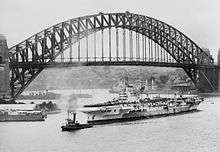 Black and white photo of a Second World War-era aircraft carrier in front of a steel through arched bridge. Several other ships are visible near the aircraft carrier