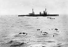 A large warship lowers boats to pick up sailors floating in the open water
